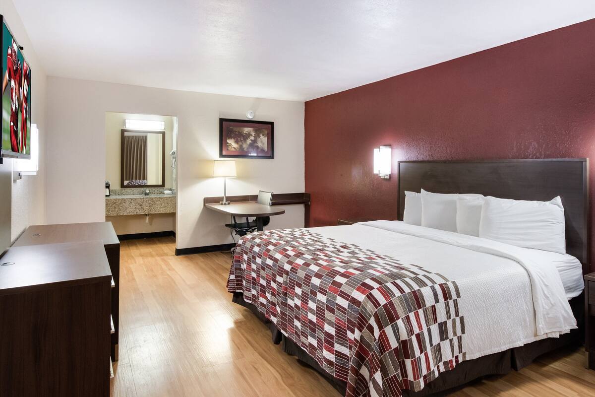 A hotel room with a bed, a desk, a mirror, a lamp, and a painting on the wall.