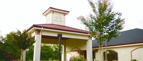 A building with a covered entrance, a tower with a clock, and a garden with various plants.