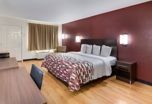 A hotel room with a bed, a desk, a chair, a nightstand, and a window with curtains. - Red Roof Inn & Suites Commerce - Athens (Commerce)
