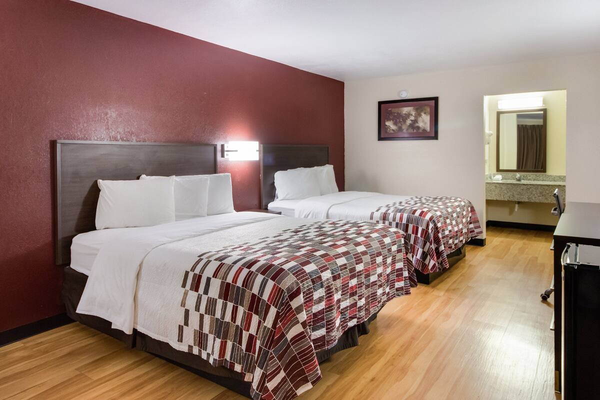 A hotel room with two beds, wooden flooring, a red accent wall, a painting, and a bathroom with a mirror and sink.