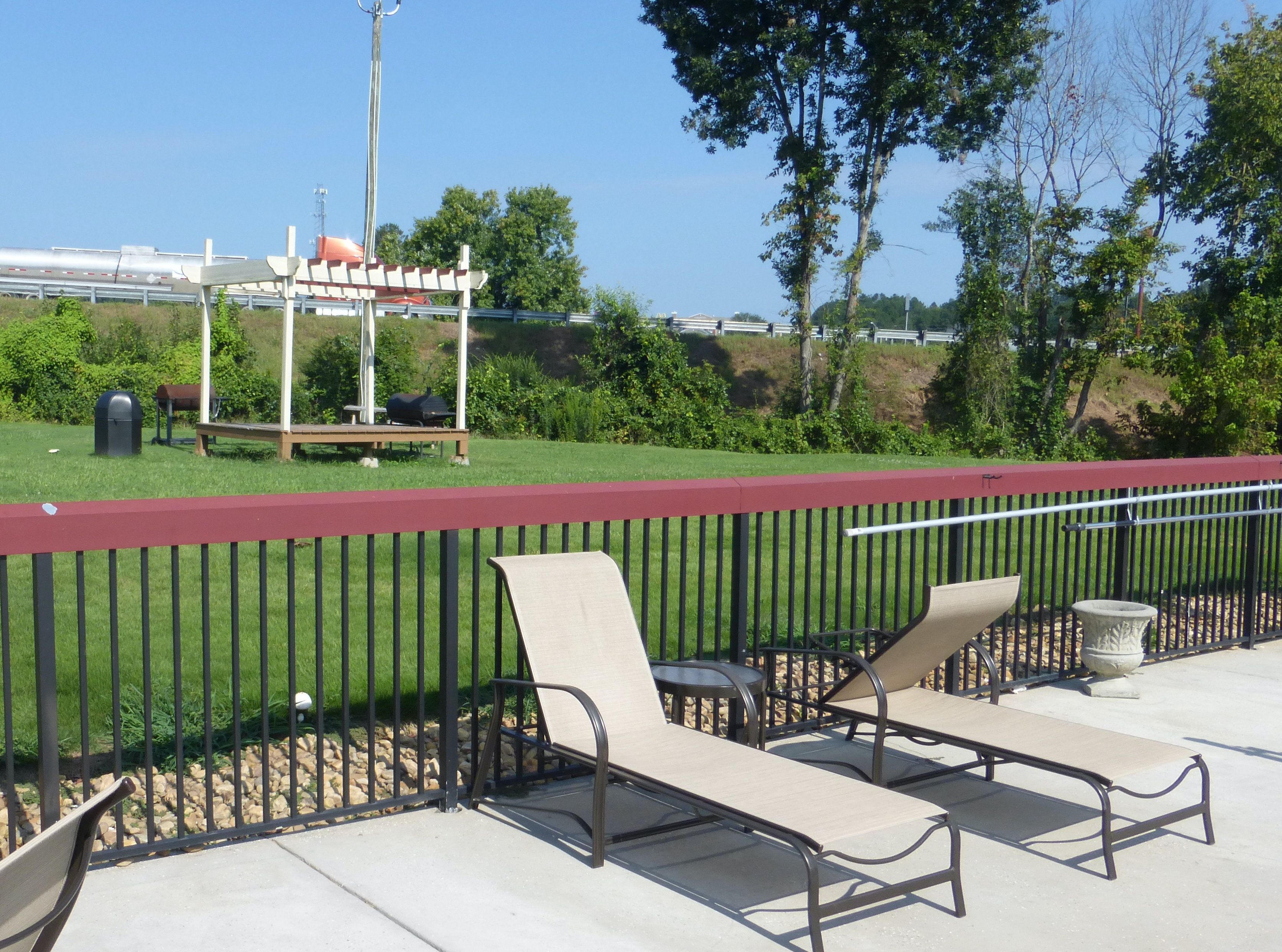 An outdoor seating area with metal chairs and a table, a red fence, and a grassy field with trees in the background.