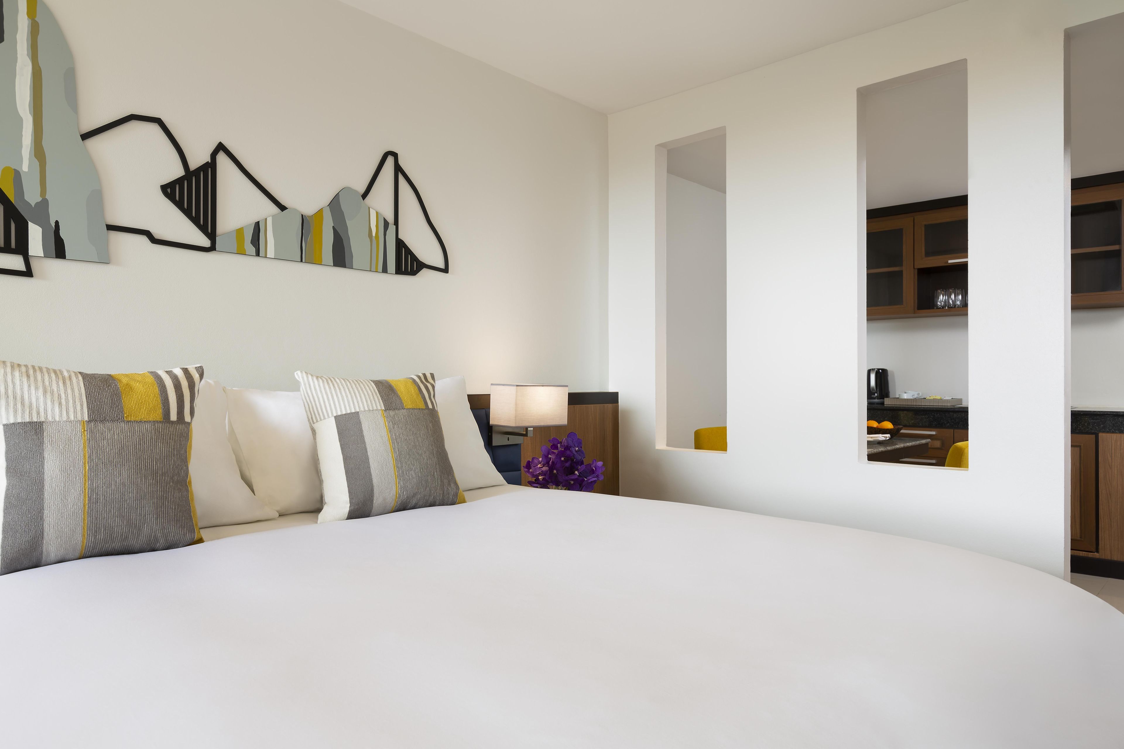 A modern hotel room with a large bed, a bedside table with a lamp, and a view into the kitchen.
