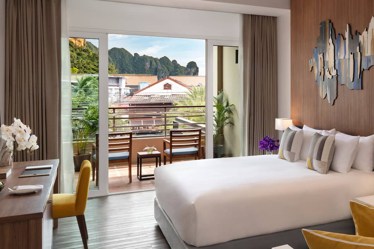 Habitación de hotel con una cama grande, un escritorio con un frutero, una silla, un balcón con vistas a edificios y follaje, y un televisor montado en la pared.