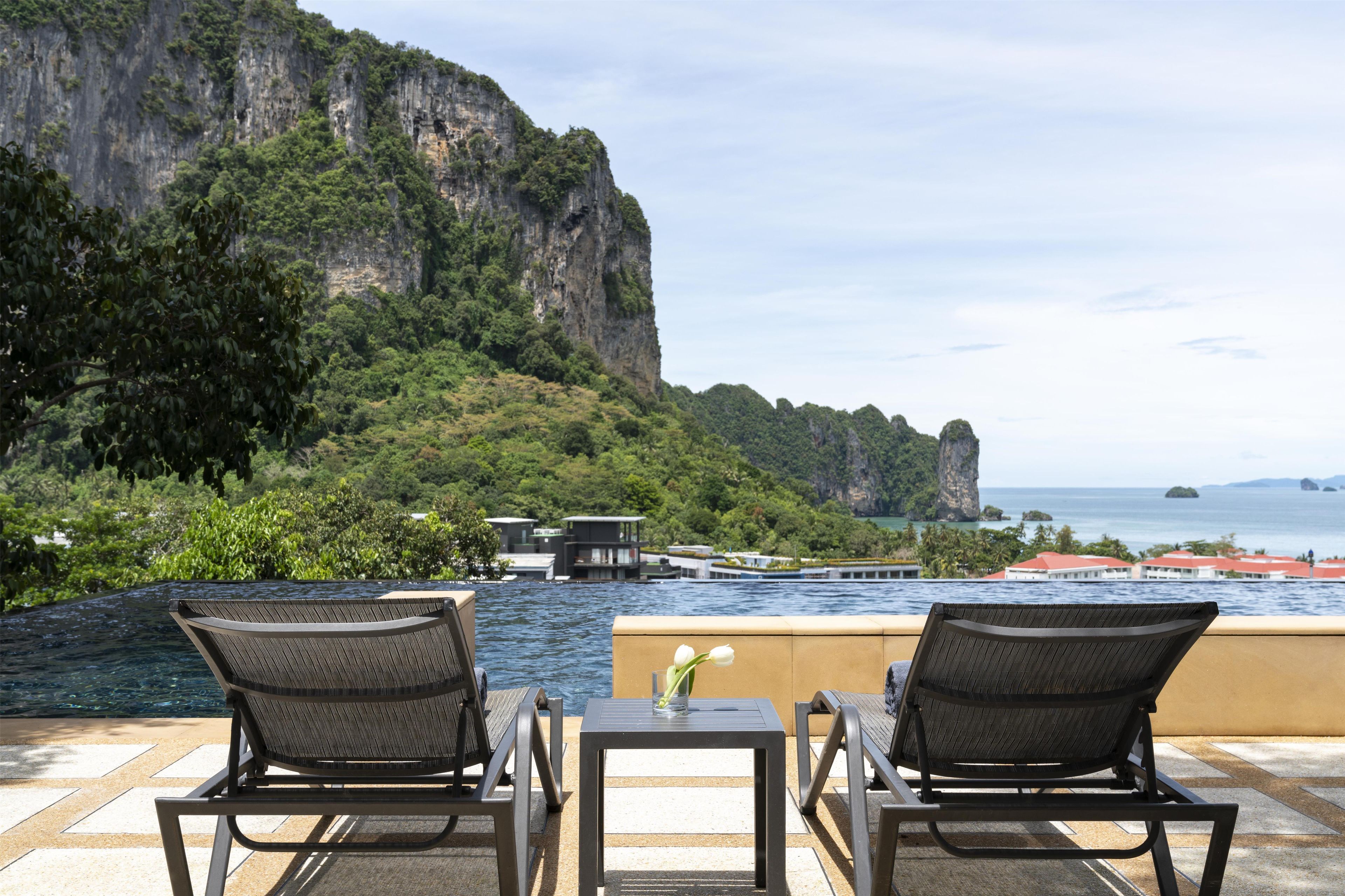 A terrace with two chairs and a table overlooking a body of water, with a cliff and buildings in the background.