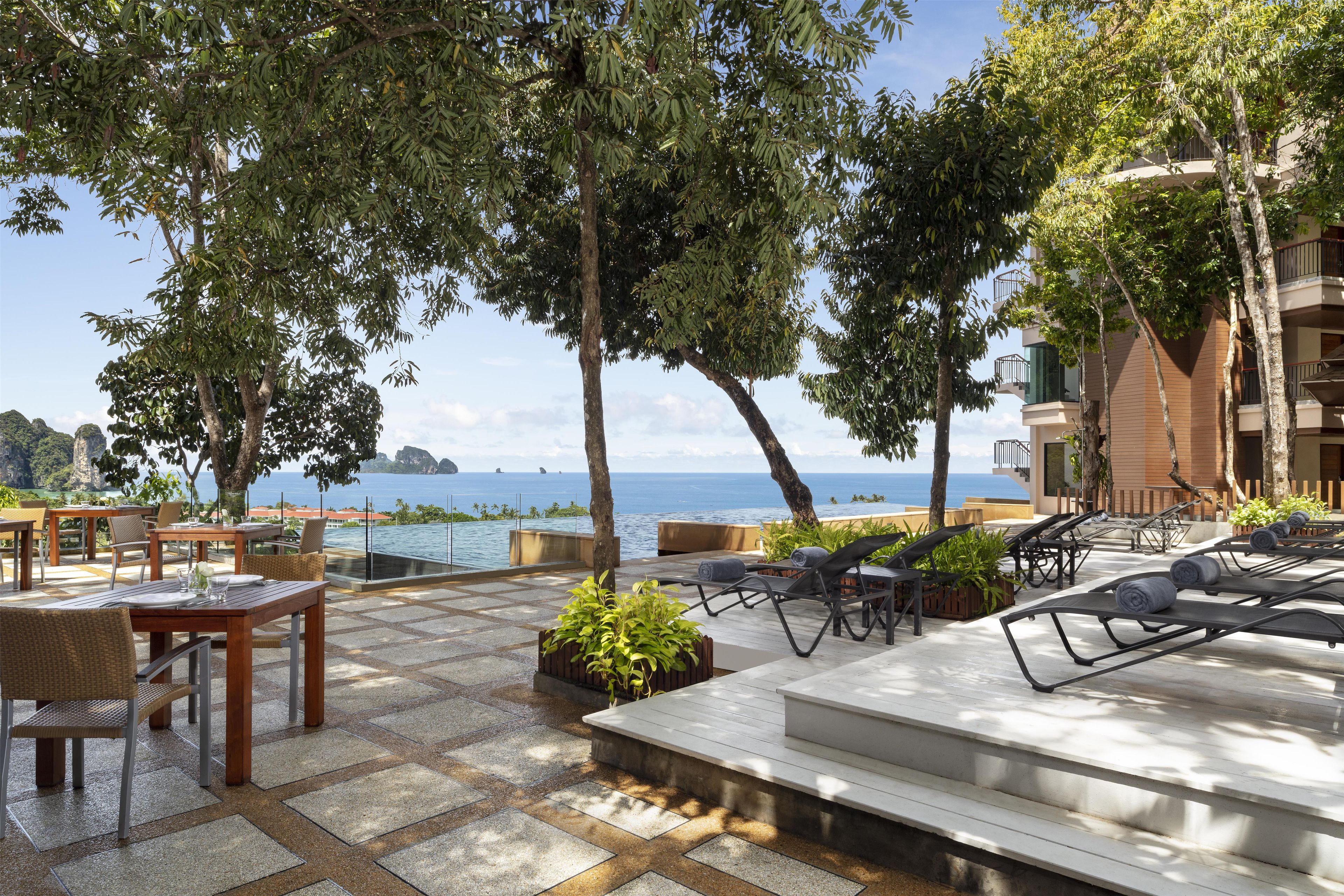 An outdoor seating area with tables and chairs, overlooking a body of water and a mountainous landscape.