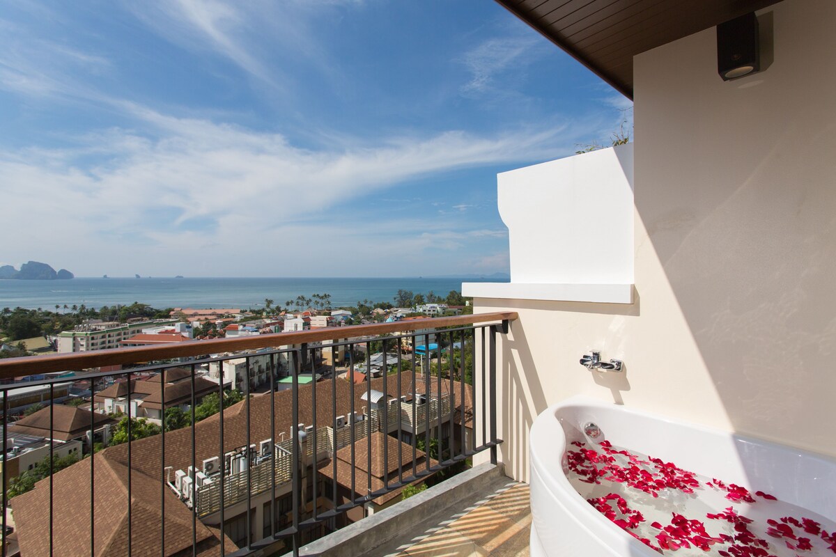 Un balcón con vista al mar, una bañera blanca con pétalos de rosa y una barandilla.