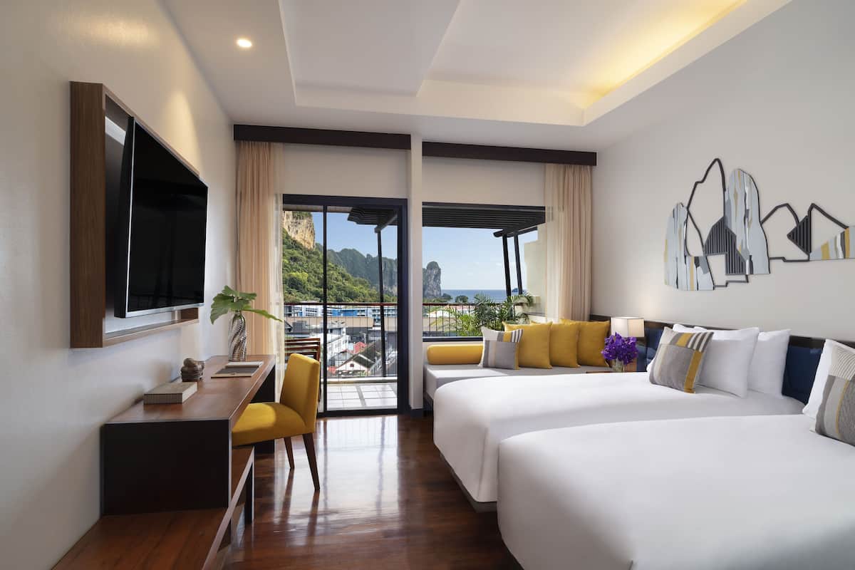 Habitación de hotel con dos camas, un escritorio y un televisor. La habitación tiene un balcón con vistas a montañas y al paisaje urbano.