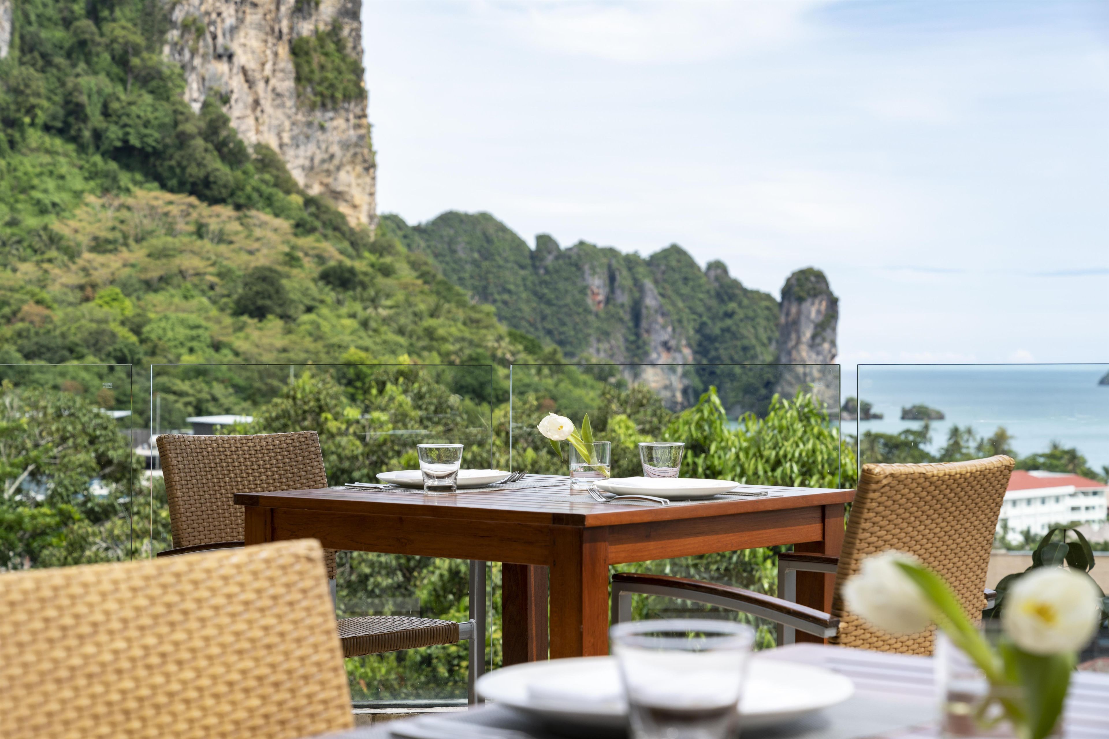 A dining table with chairs, a glass of water, and a flower, set against a backdrop of a cliff and ocean view.