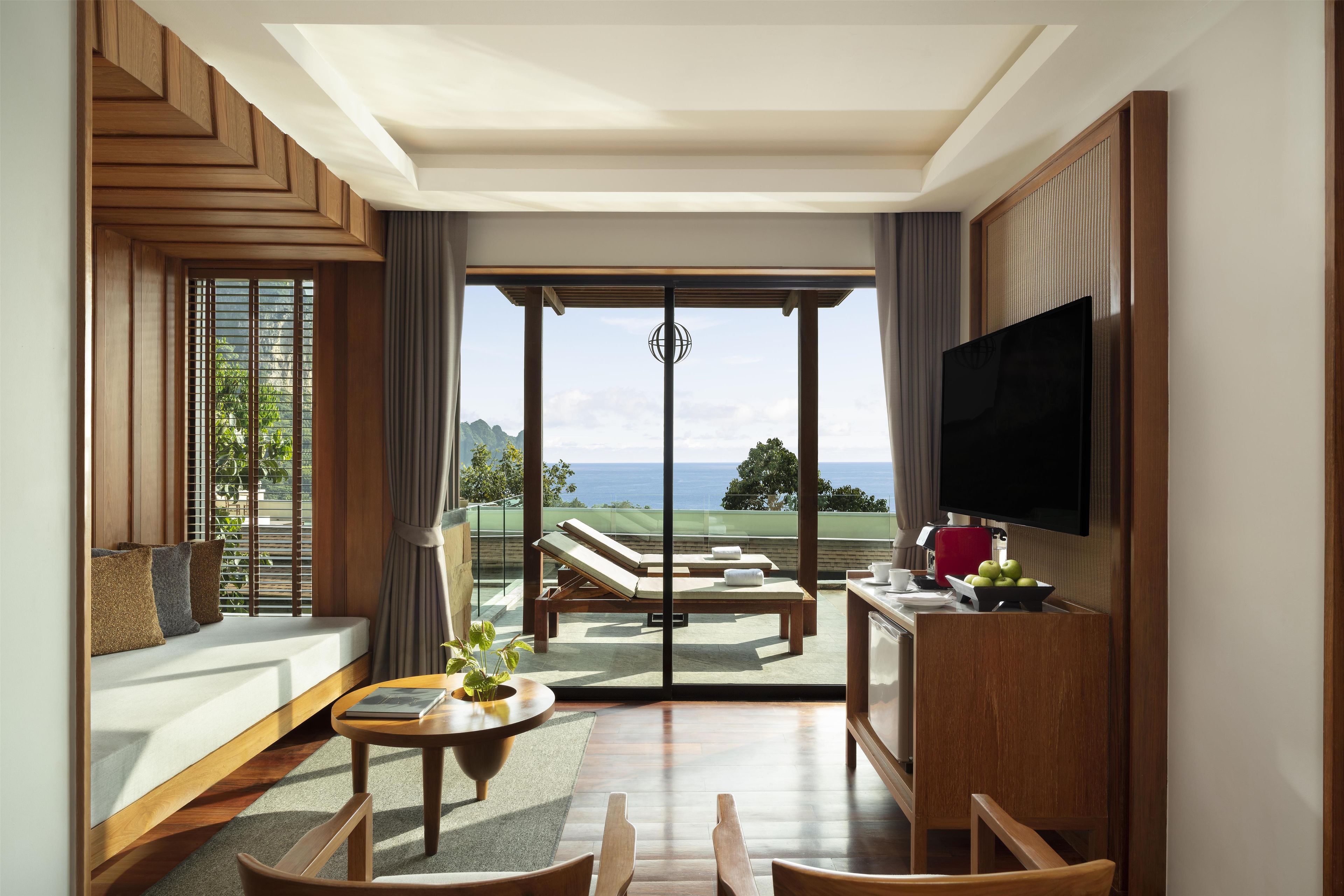 A spacious living area with a large sliding glass door leading to a balcony with a view of the ocean. There is a flat-screen TV mounted on the wall, a wooden coffee table, and a seating area with a sofa and chairs.
