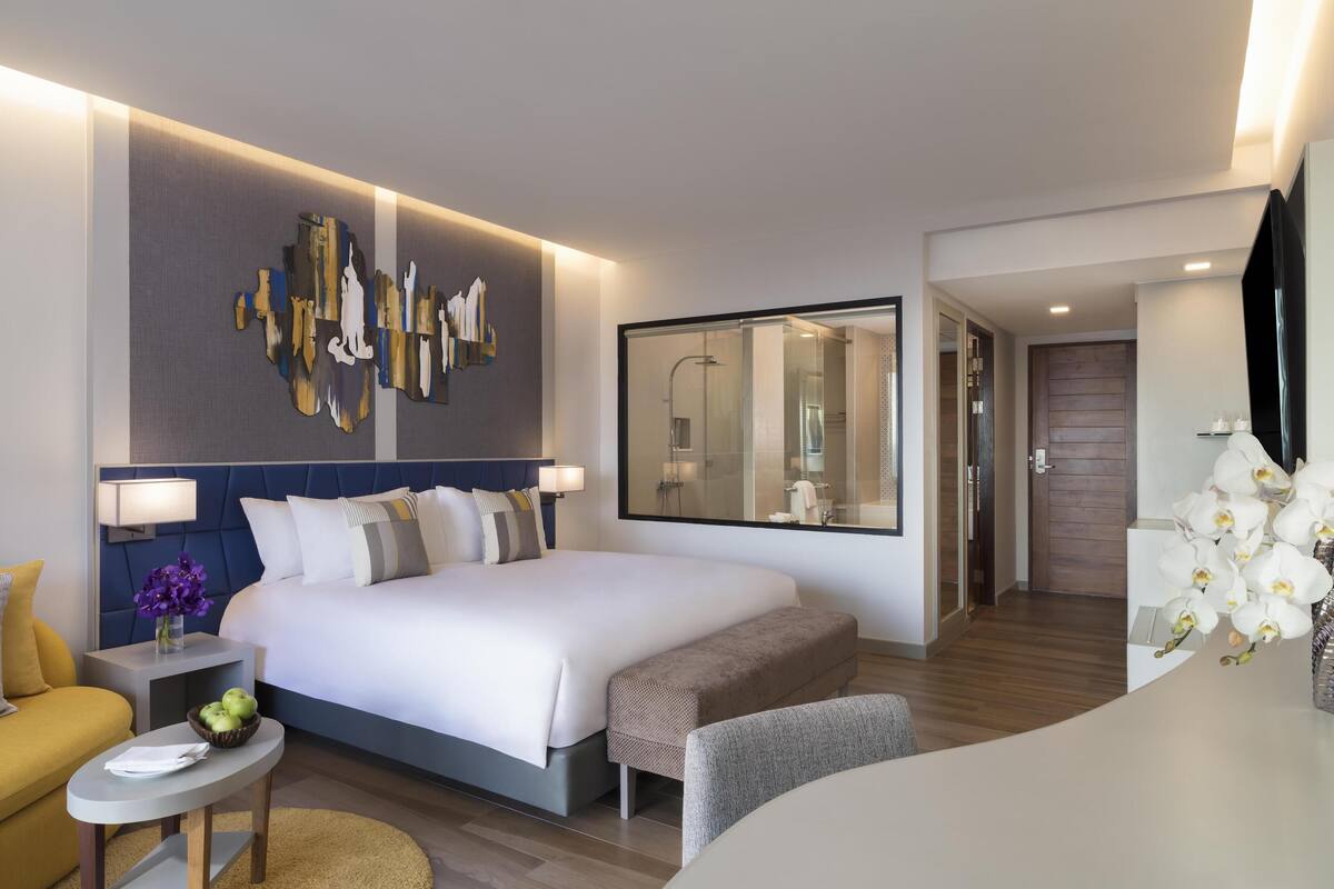 Una habitación de hotel moderna con una cama grande, una mesita de noche con lámpara, una pequeña mesa redonda con un frutero, un banco y un espejo grande.