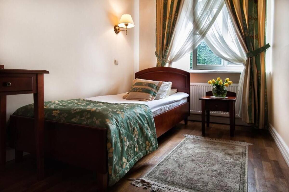 A bedroom with a wooden bed, a bedside table with a lamp, a window with sheer curtains, and a floral-patterned bedspread.