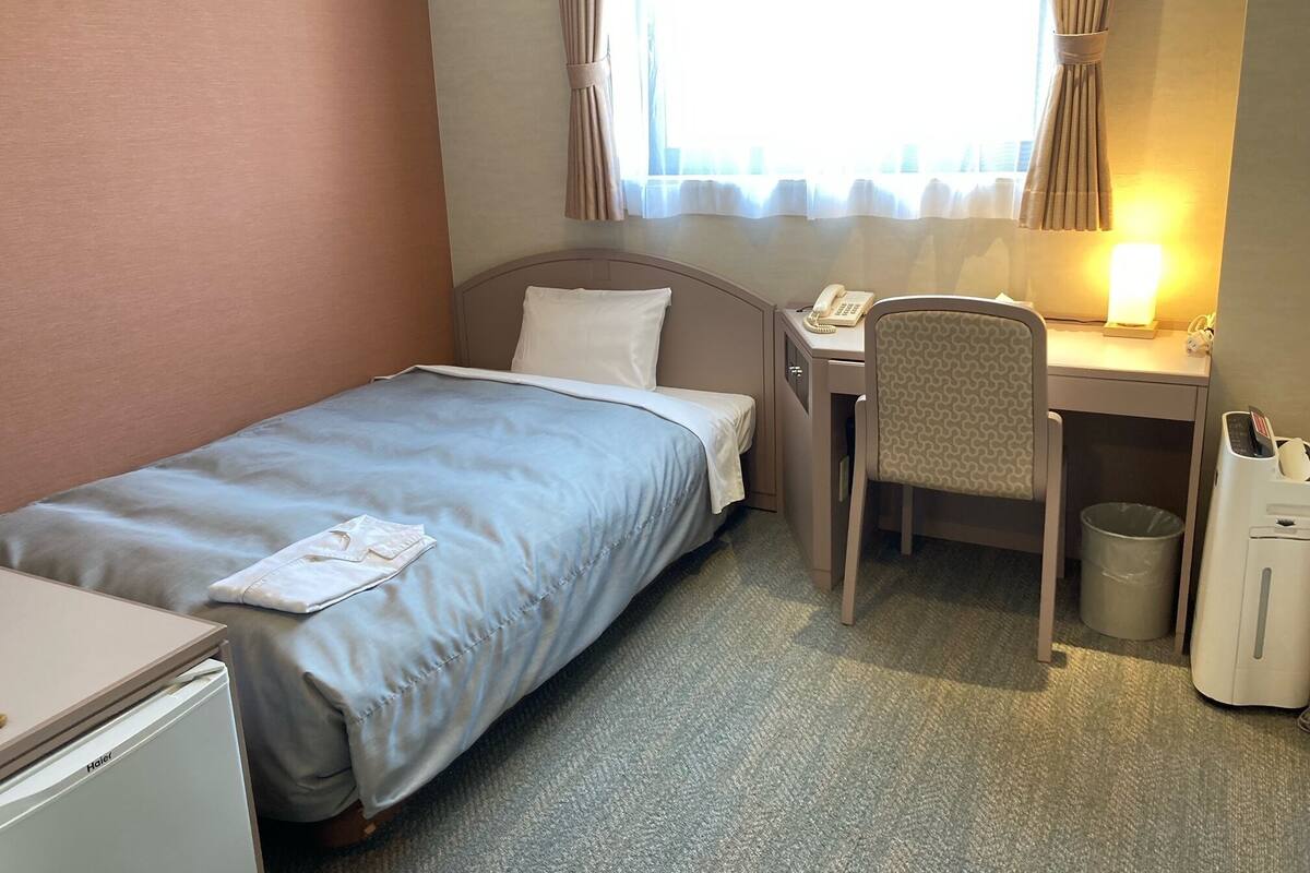 Quarto de hotel com uma cama de solteiro, uma escrivaninha com cadeira, uma luminária de mesa, um telefone, um cesto de lixo e um aquecedor.