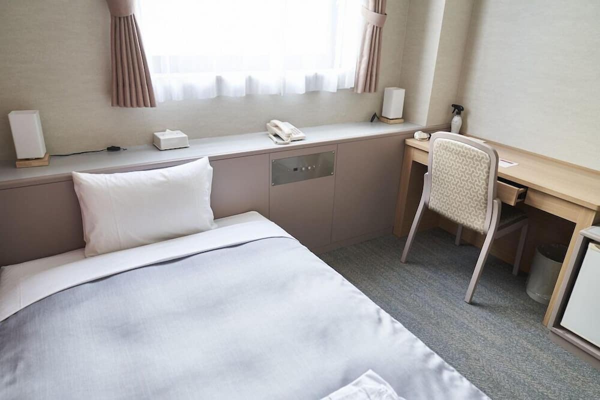 Quarto de hotel com cama, escrivaninha, cadeira, telefone e janela com cortinas.
