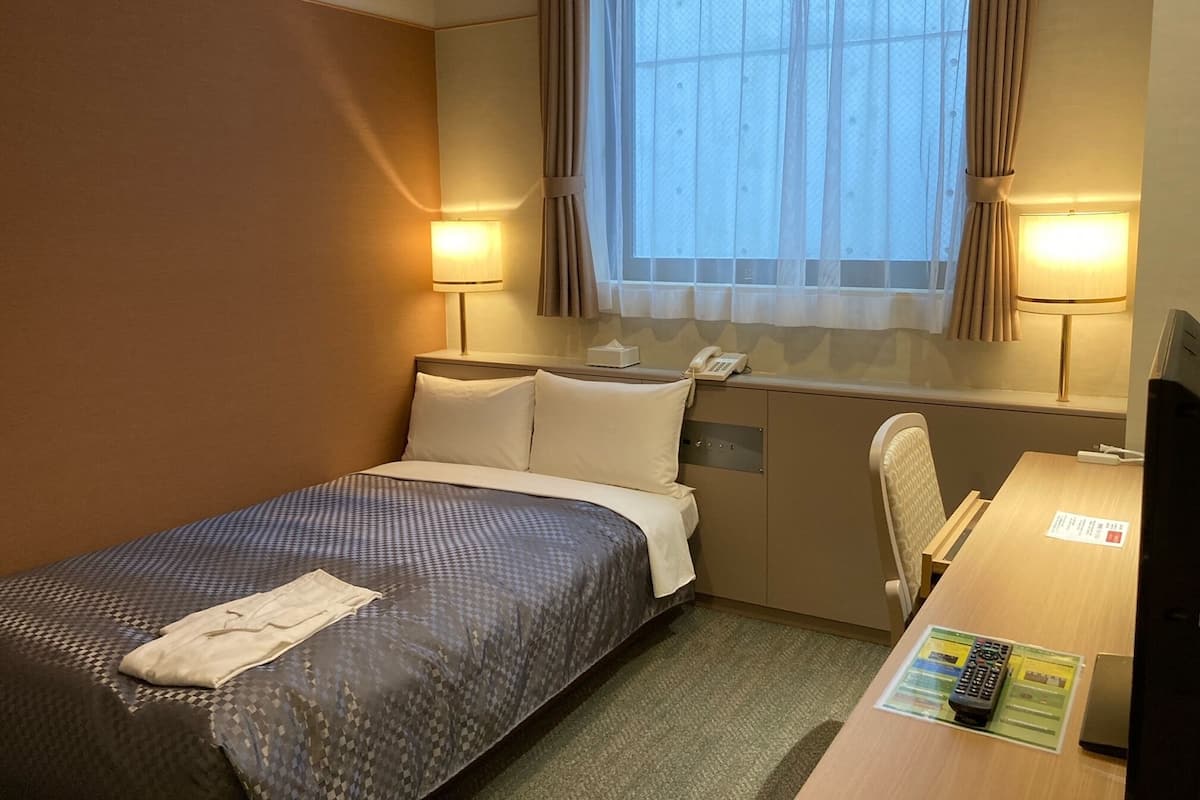 Quarto de hotel com uma cama de solteiro, uma mesa com computador, telefone e janela com cortinas.