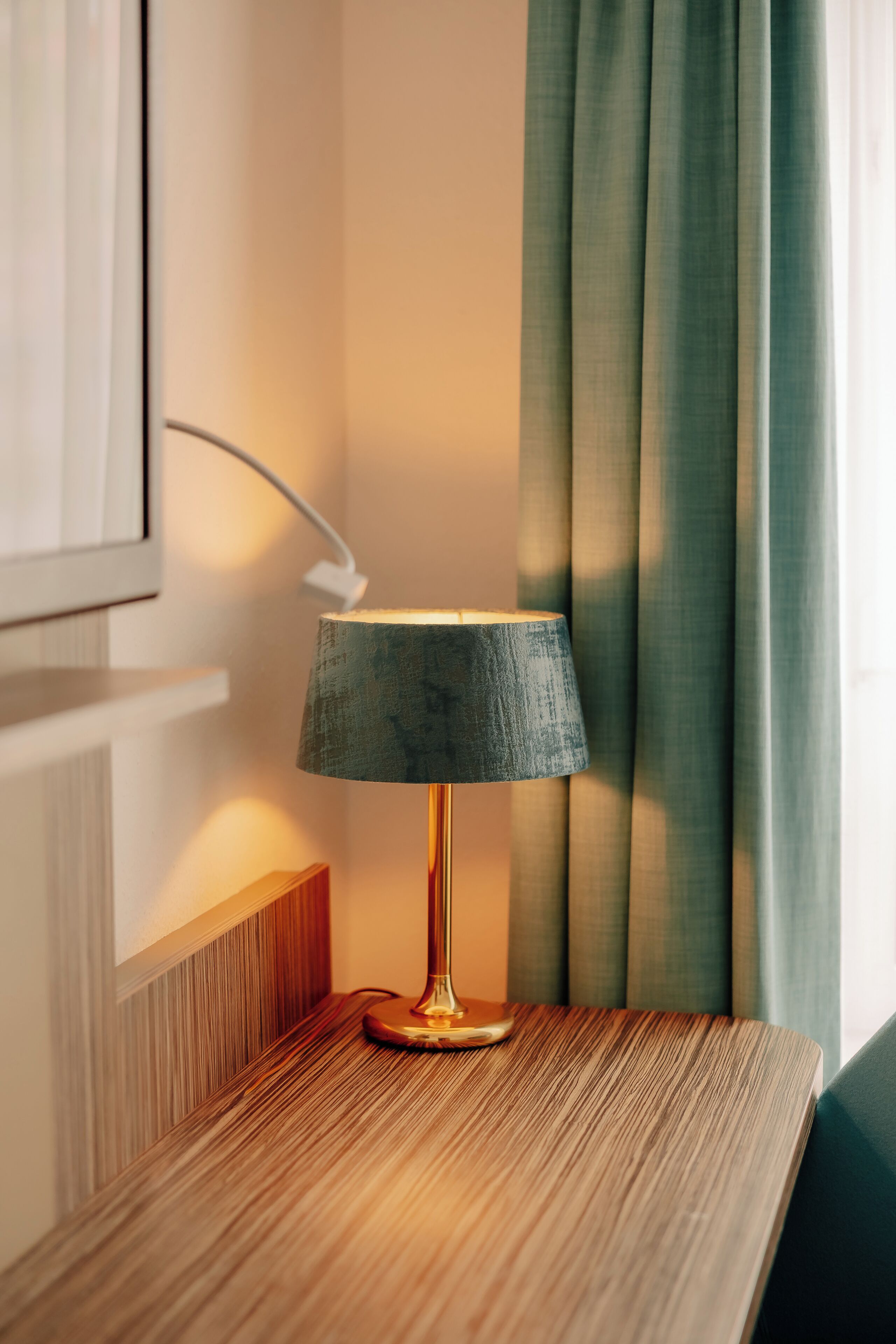 A lamp on a wooden bedside table with a power cord plugged in, a wall-mounted picture frame, and a green curtain partially drawn.