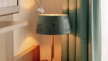 En lampe på et trænatbord med en tilsluttet ledning, en billedramme hængt på væggen og en grøn gardin, der er halvt trukket fra.