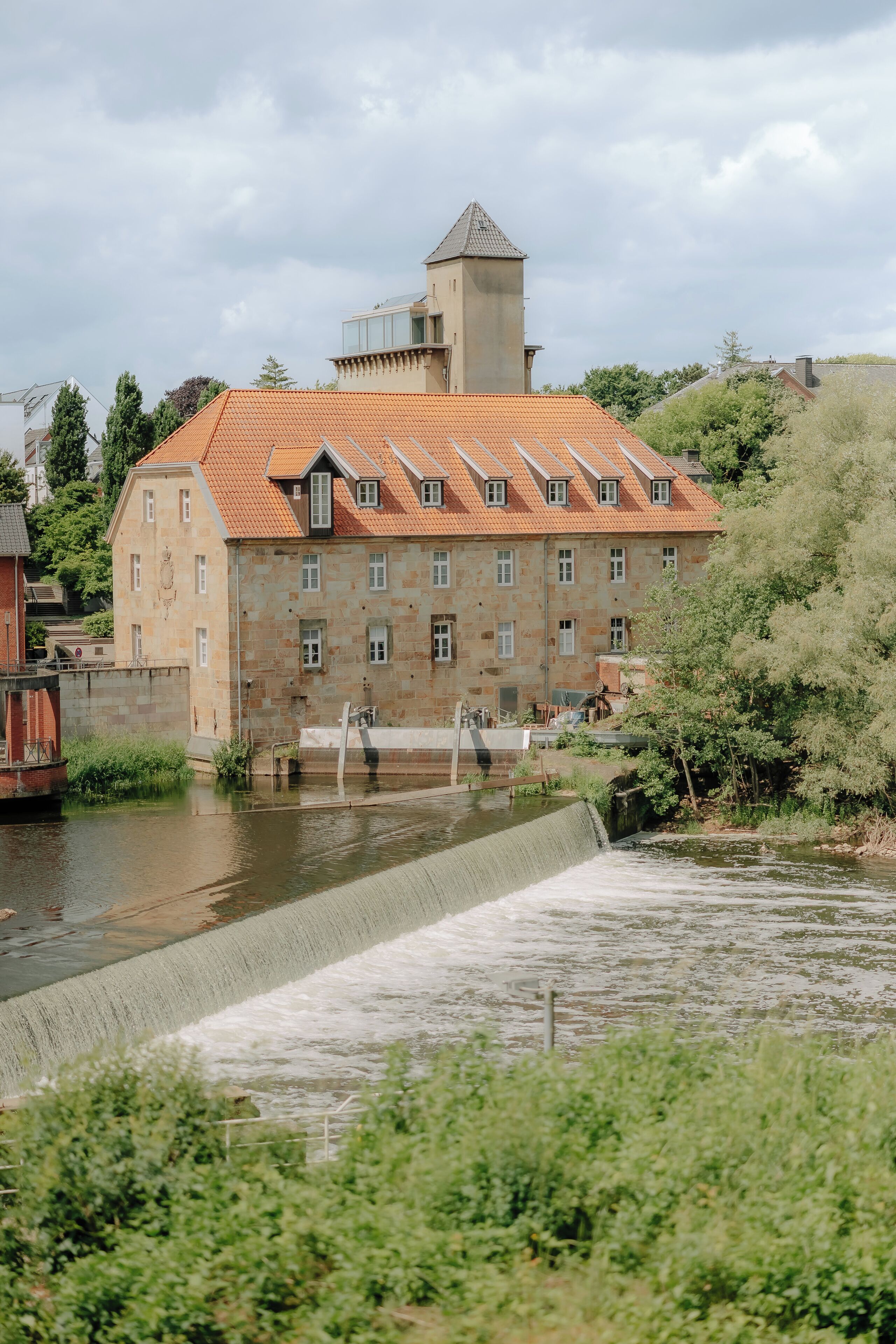 A historic building with a stone facade and a red-tiled roof, situated beside a water channel.