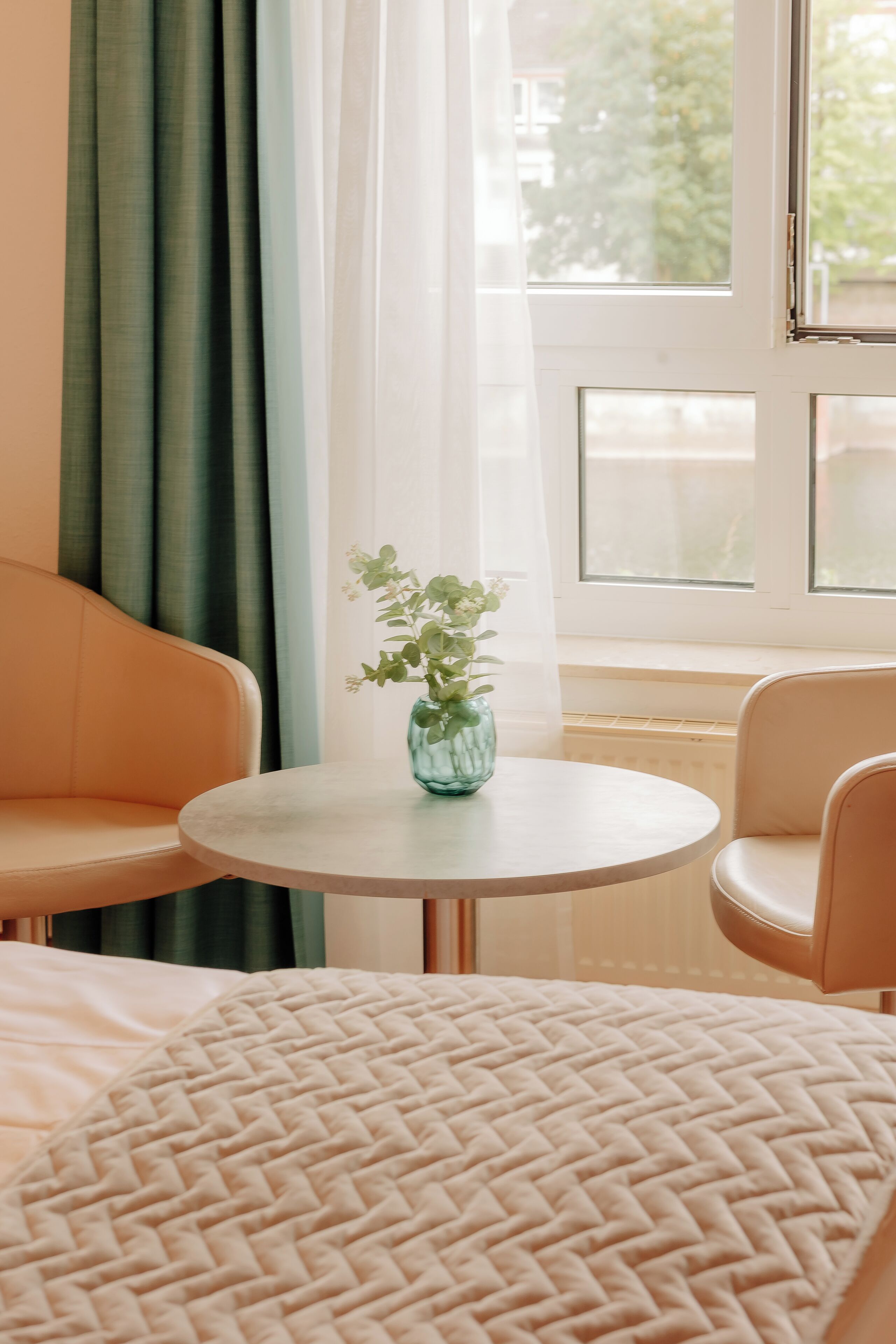 A room with a bed, a round table with a vase, a sofa, and a window with curtains.