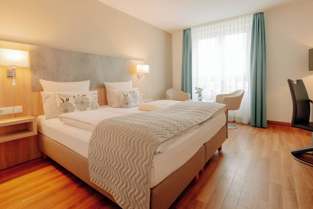 A hotel room with a large bed, two bedside tables, a chair, a desk, and a window with curtains.