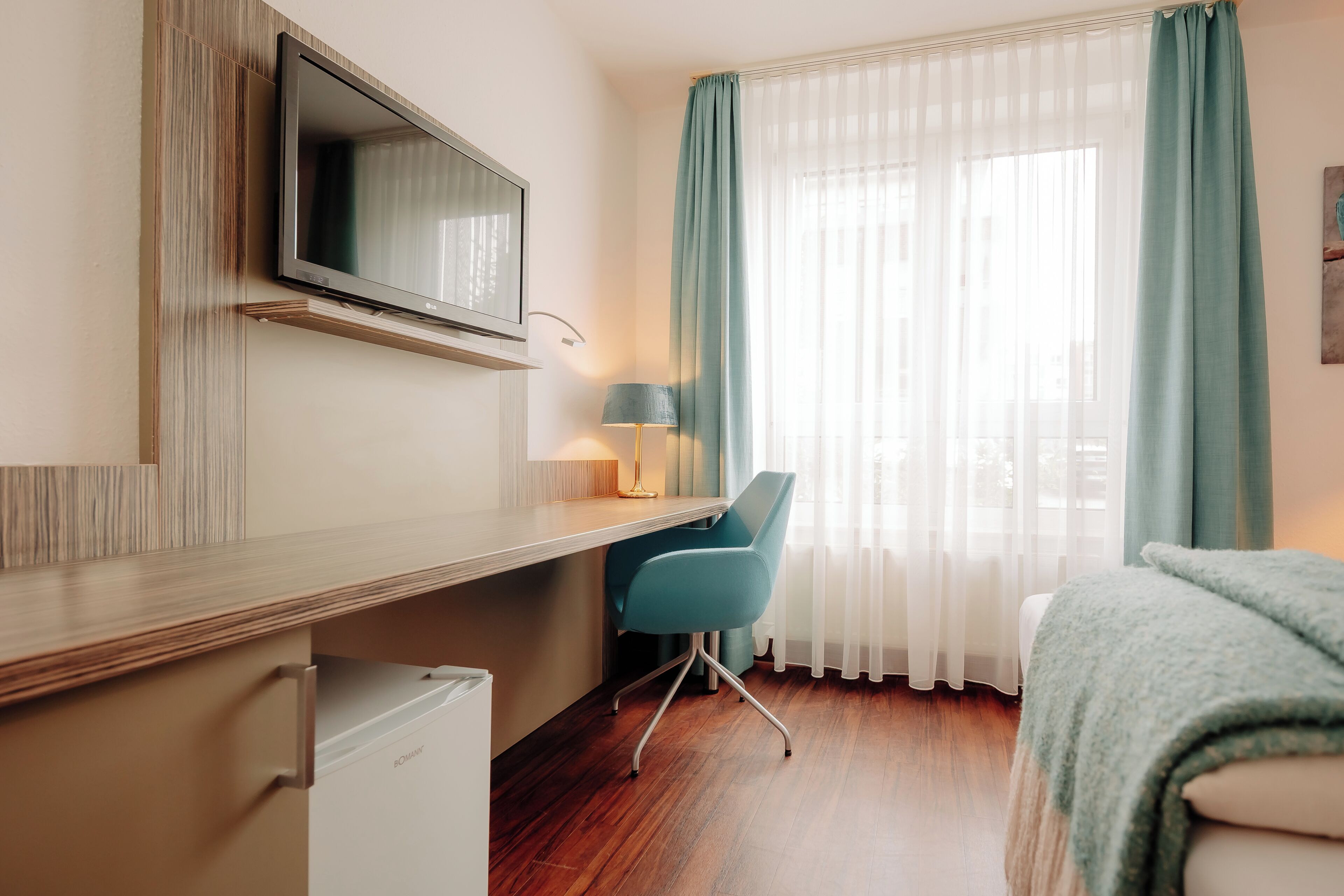 A hotel room with a wooden desk, a television, a chair, a bed, and a window with curtains.