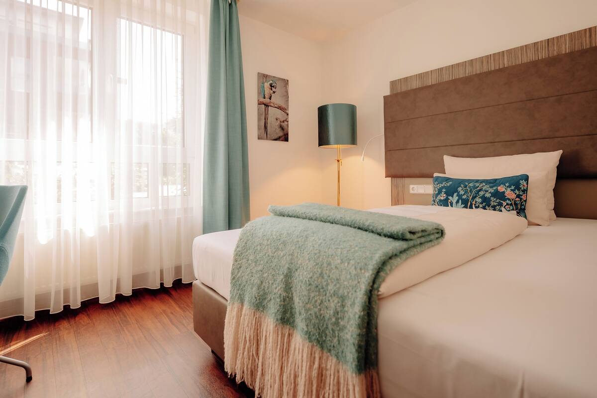 A hotel room with a bed, a chair, a lamp, and a window with curtains.