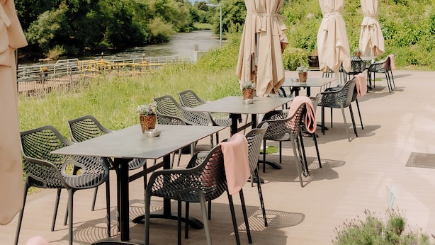 An outdoor patio with tables and chairs, beige umbrellas, and a view of a river and greenery. - CASCADA Rheine by Hackmann (Rheine)