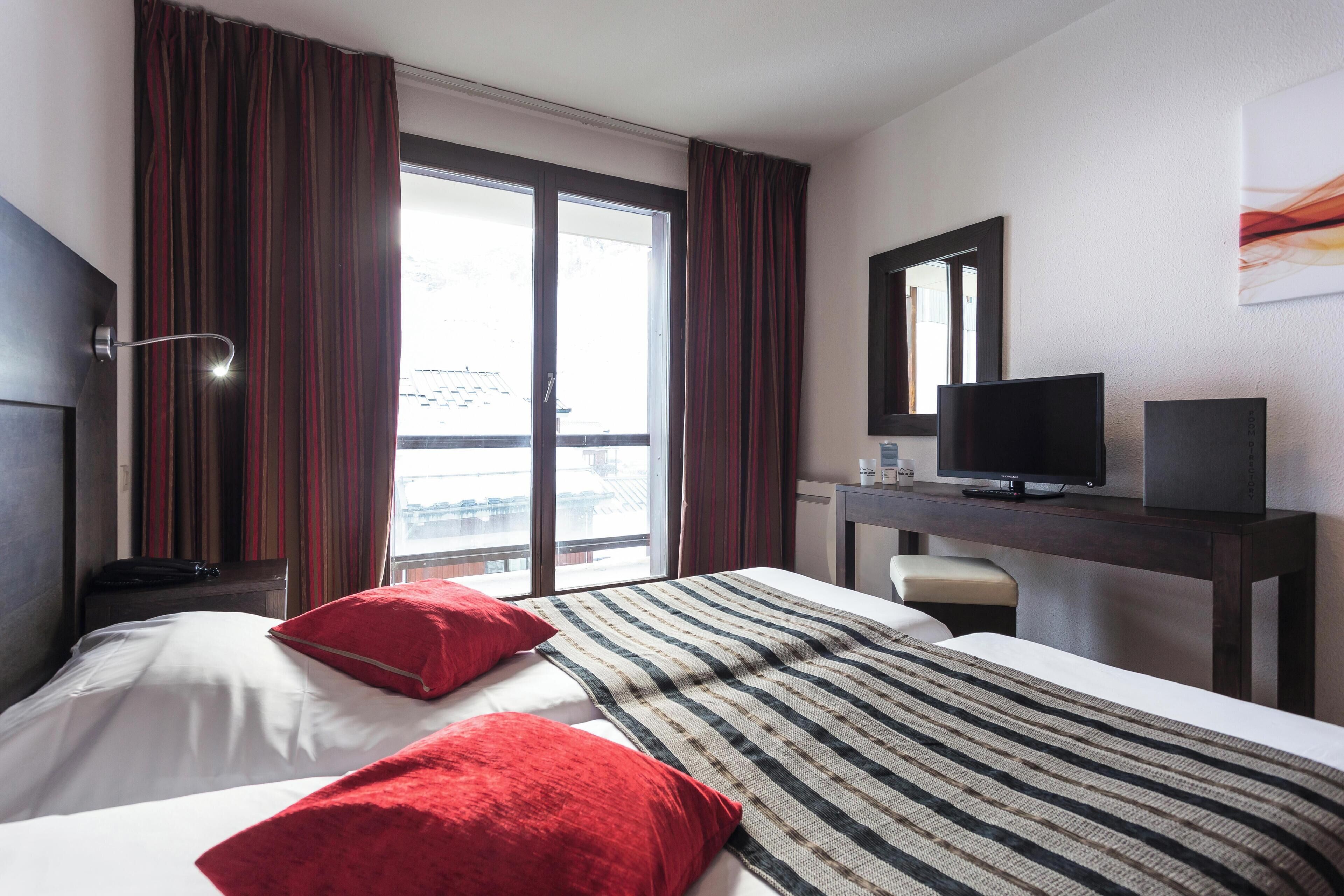 A hotel room with two beds, a desk with a TV, and a large window with curtains.