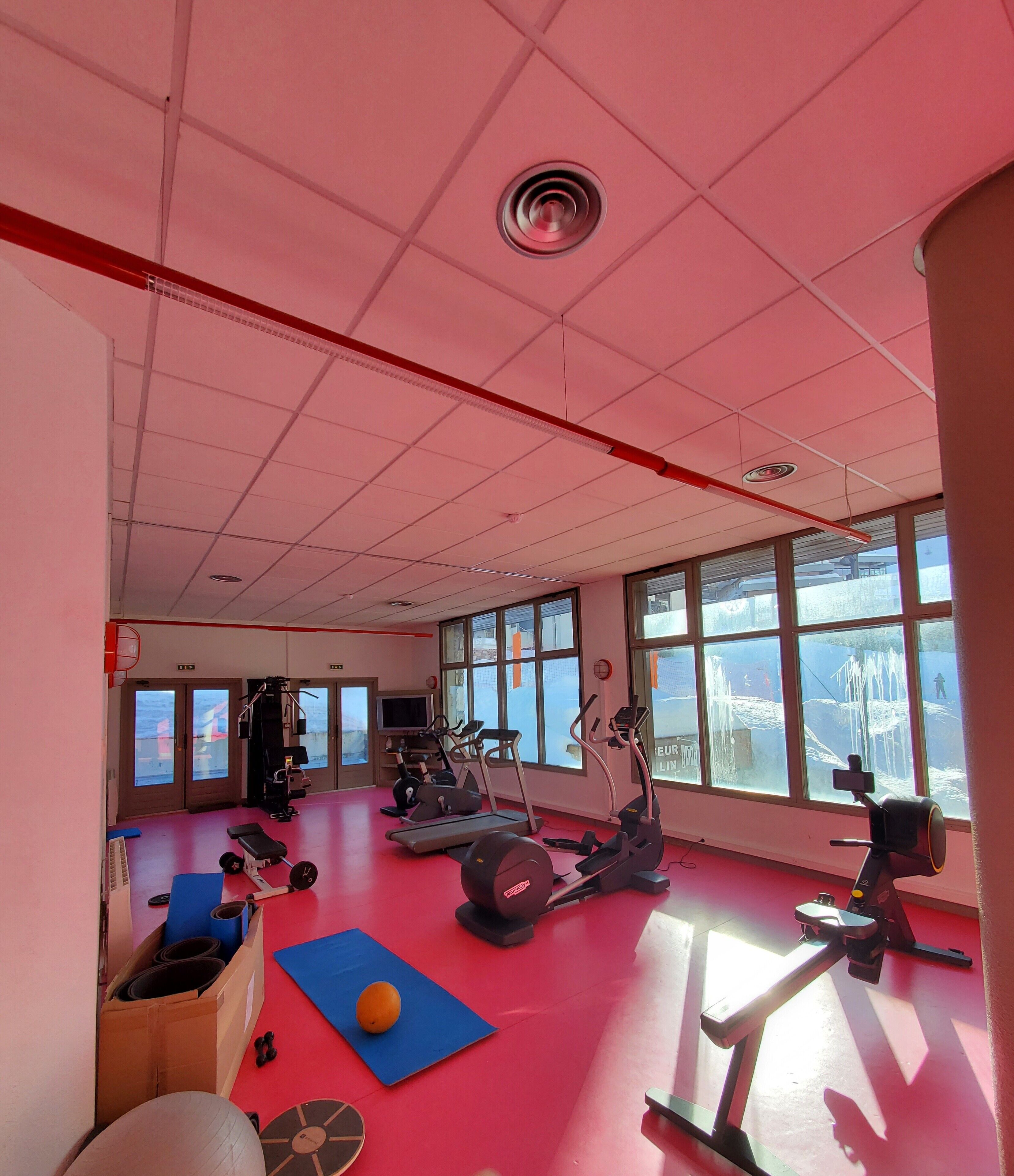 Et fitnesscenter med forskellig træningsudstyr, store vinduer og rødlakeret gulv.