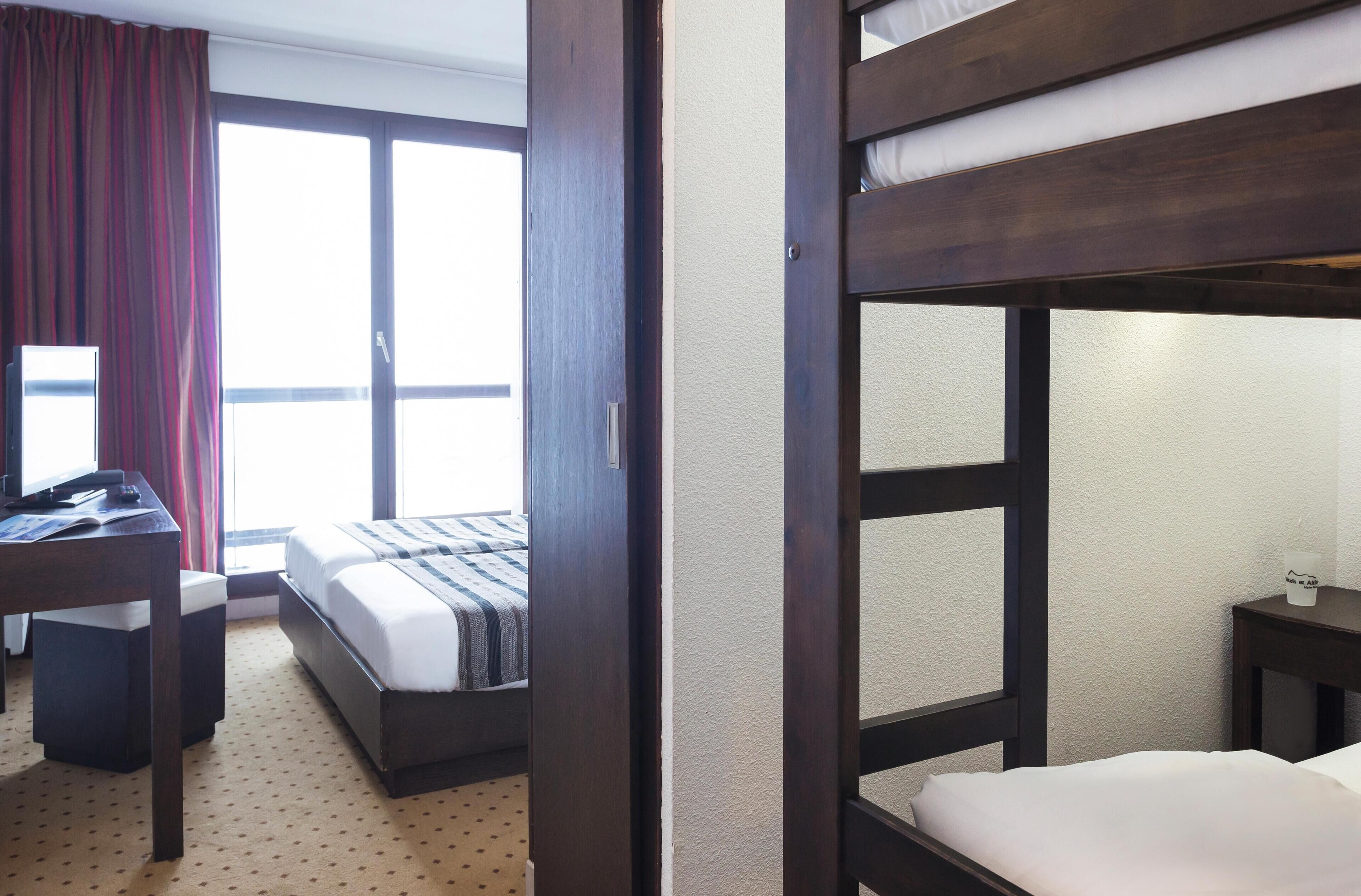 Una habitación de hotel con literas, un escritorio y una silla, una mesita pequeña y una ventana con cortinas.