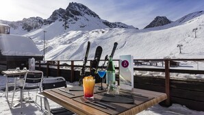 Una mesa de madera con una bebida, esquíes y un cartel en un entorno nevado al aire libre, con montañas de fondo.