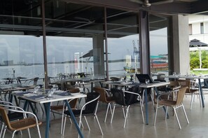 An outdoor dining area with tables and chairs, large windows, and a view of the water and buildings. - Xandari Harbour Fort Kochi (Kochi)