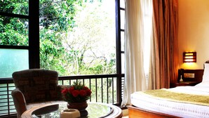 A room with a bed, a chair, a table with a vase of flowers, and a view of greenery through the window.