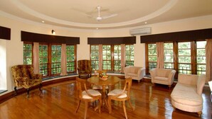 A spacious room with a round dining table, several chairs, and a ceiling fan. Large windows with curtains provide natural light and views of greenery outside.