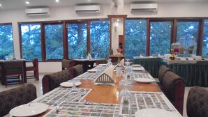 A dining area with a long table set for a meal, surrounded by chairs, with large windows offering a view of trees.