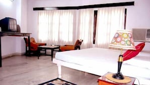 A hotel room with a bed, a desk with a television, two chairs, a lamp, and a window with curtains. - Hotel Mewar Inn (Udaipur)