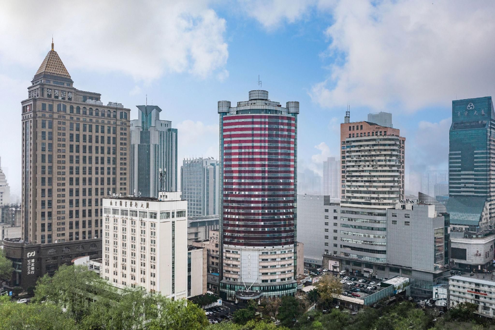 Vista urbana com diversos edifícios altos, incluindo um imponente arranha-céu cilíndrico com padrão vermelho e branco, cercado por outras construções modernas.