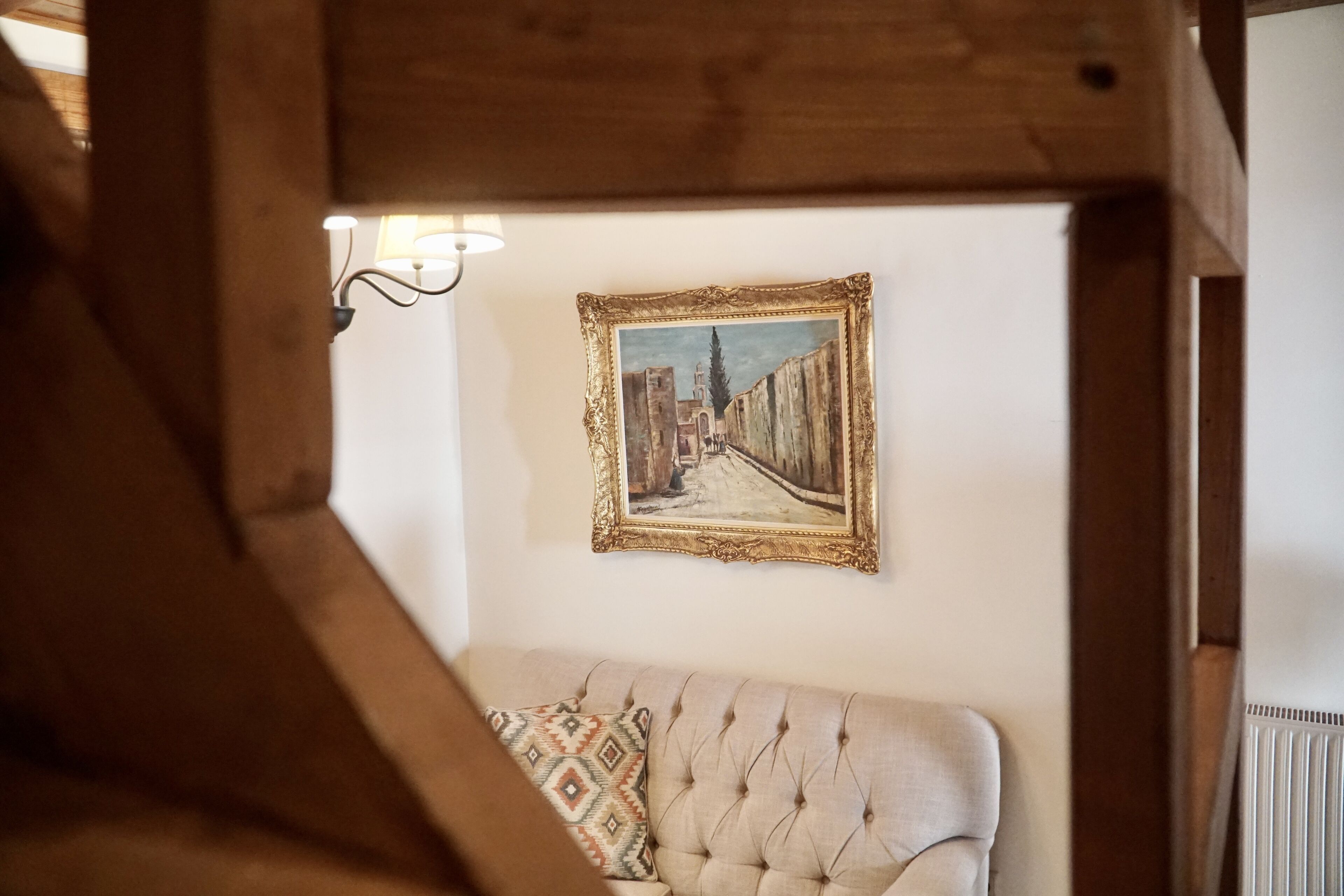 A room with a beige sofa, a framed painting of a city street, and a wall-mounted light fixture.