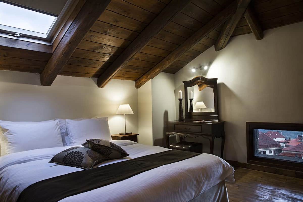 Une chambre avec une lucarne, un lit recouvert de draps blancs, une table de chevet avec une lampe, une commode avec un miroir et un téléviseur fixé au mur.