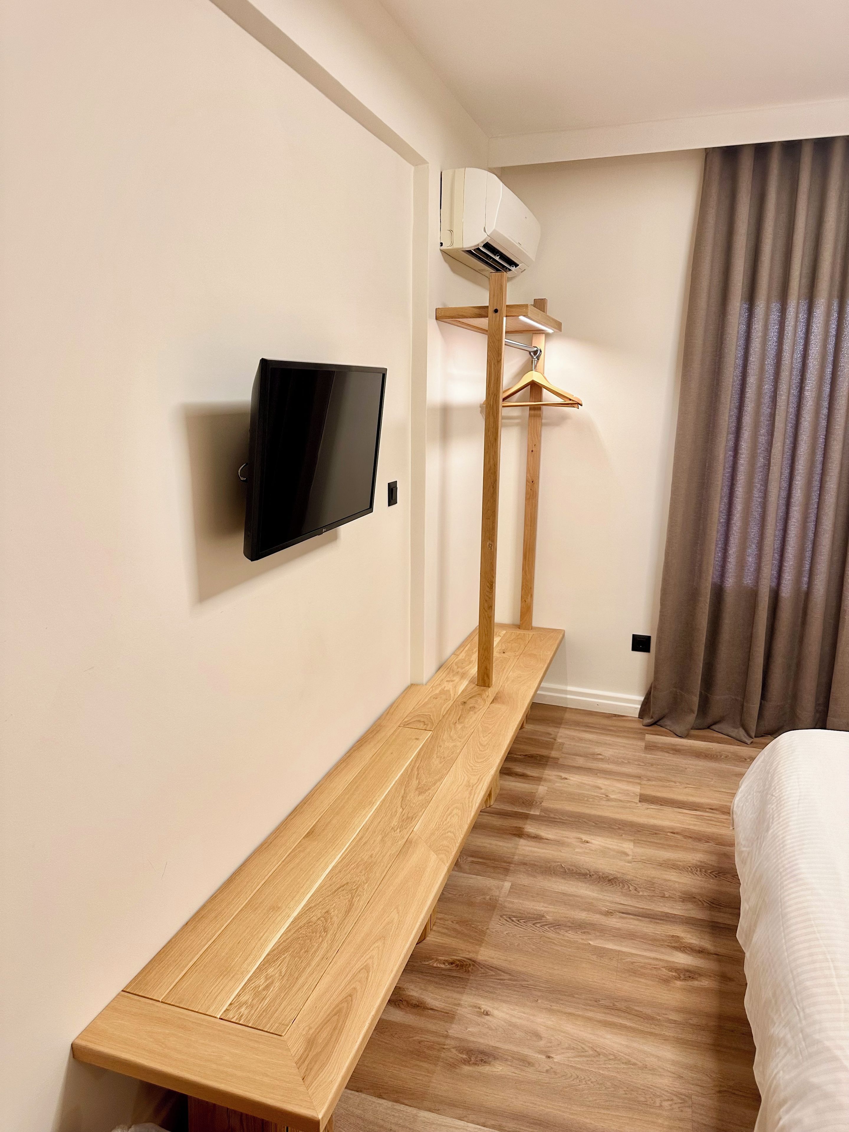 A hotel room with a wooden bench, a mounted TV, an air conditioning unit, and a bed.