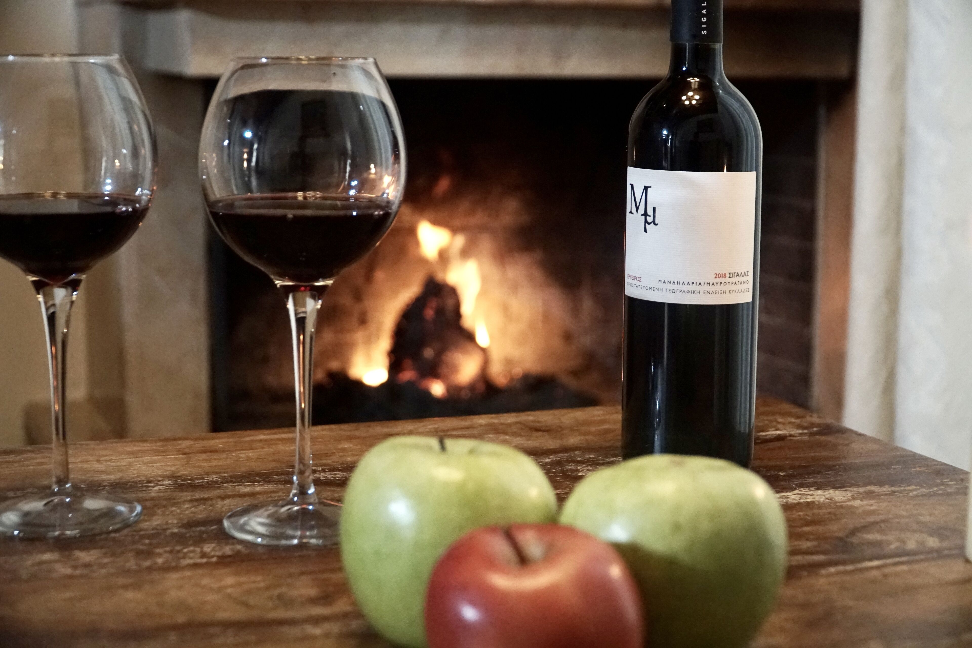 A wooden table with two wine glasses, a bottle of wine, and three apples.