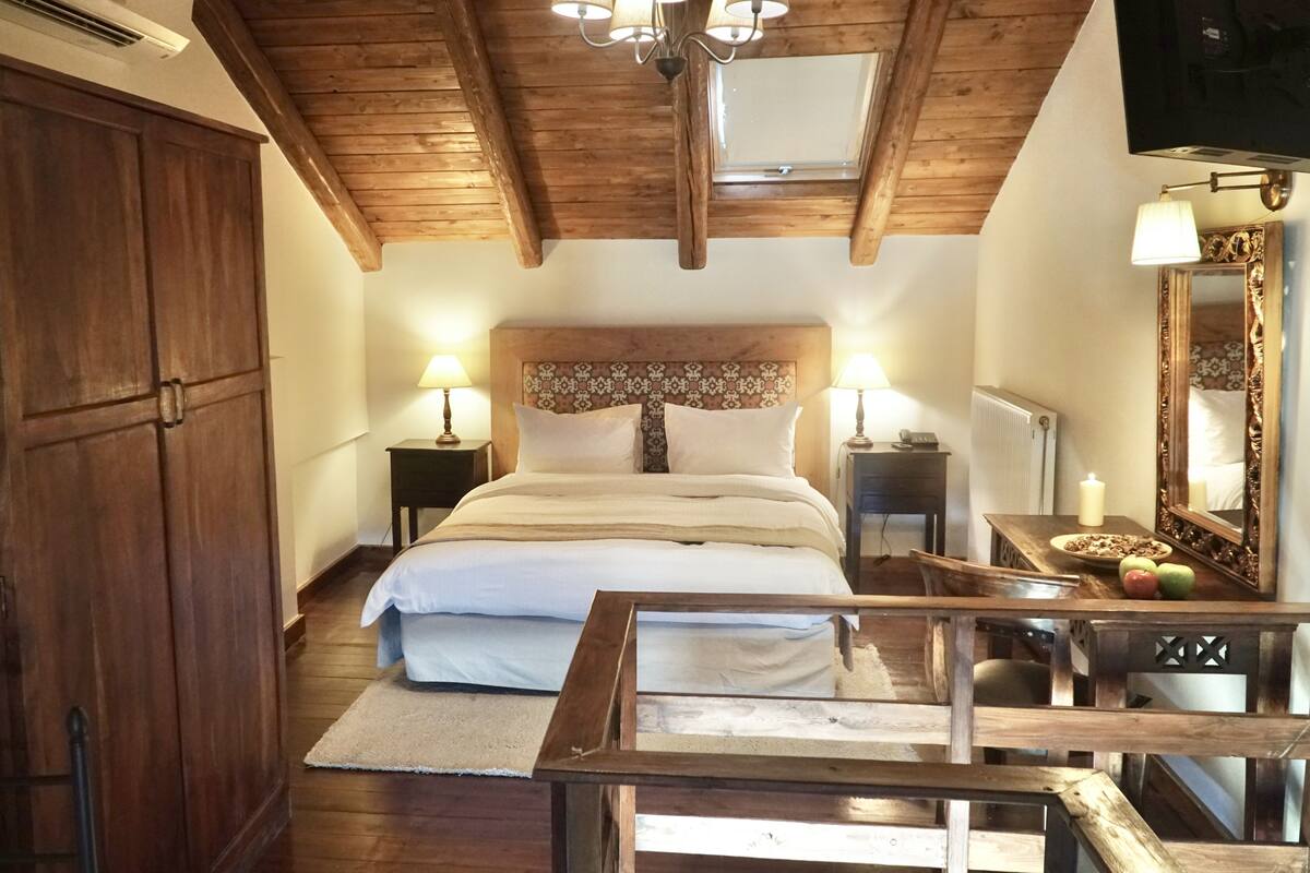 Une chambre avec un plafond en bois, un grand lit recouvert de draps blancs, deux lampes de chevet, une table de chevet en bois avec un miroir, et une petite table avec des fruits et une bougie.