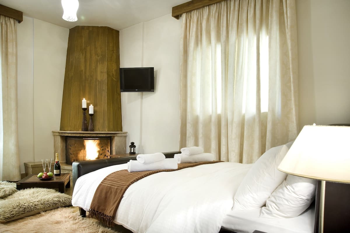 Une chambre d’hôtel avec une cheminée, un lit avec du linge de maison blanc, un téléviseur à écran plat et une table de chevet avec une bouteille de vin et des fruits.