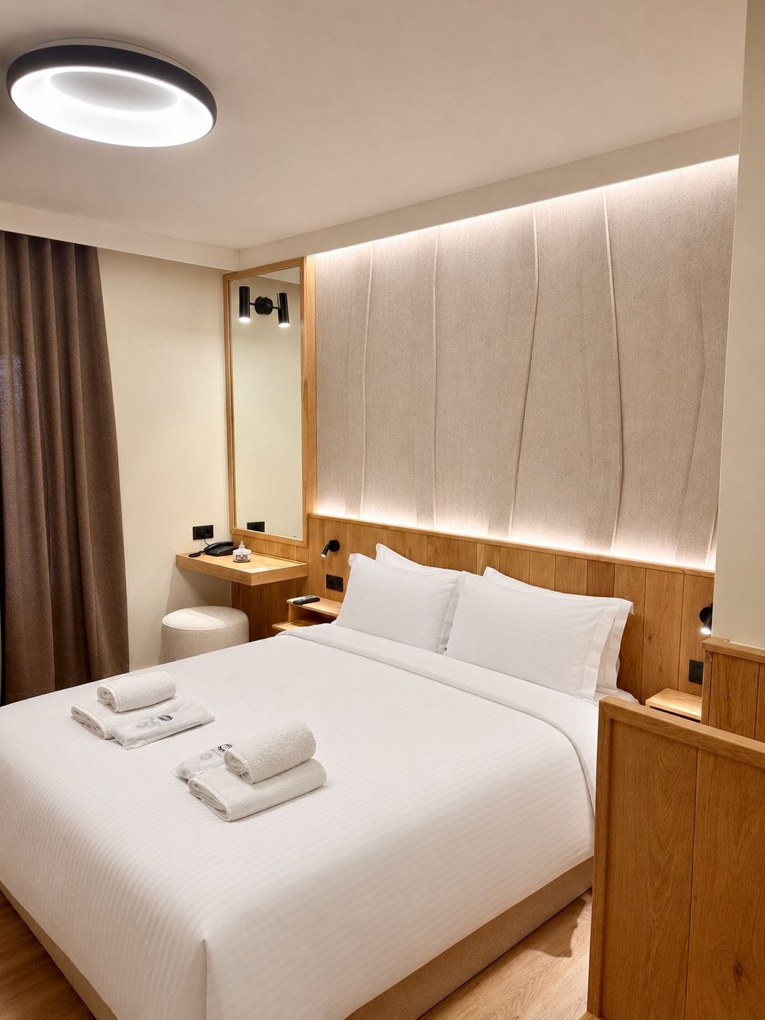 A hotel room with a large bed, a wooden headboard, a bedside table, a nightstand, a mirror, a wooden desk, a chair, and a round ceiling light.