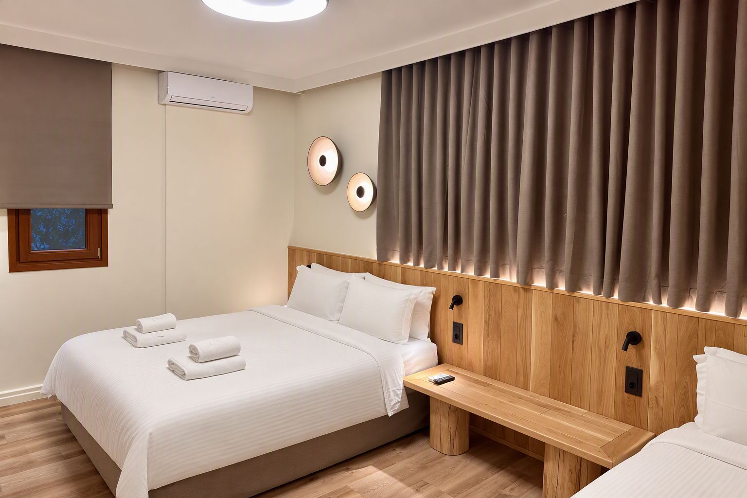 A modern hotel room with a large bed, bedside tables, a wall-mounted air conditioner, and a window with a view.