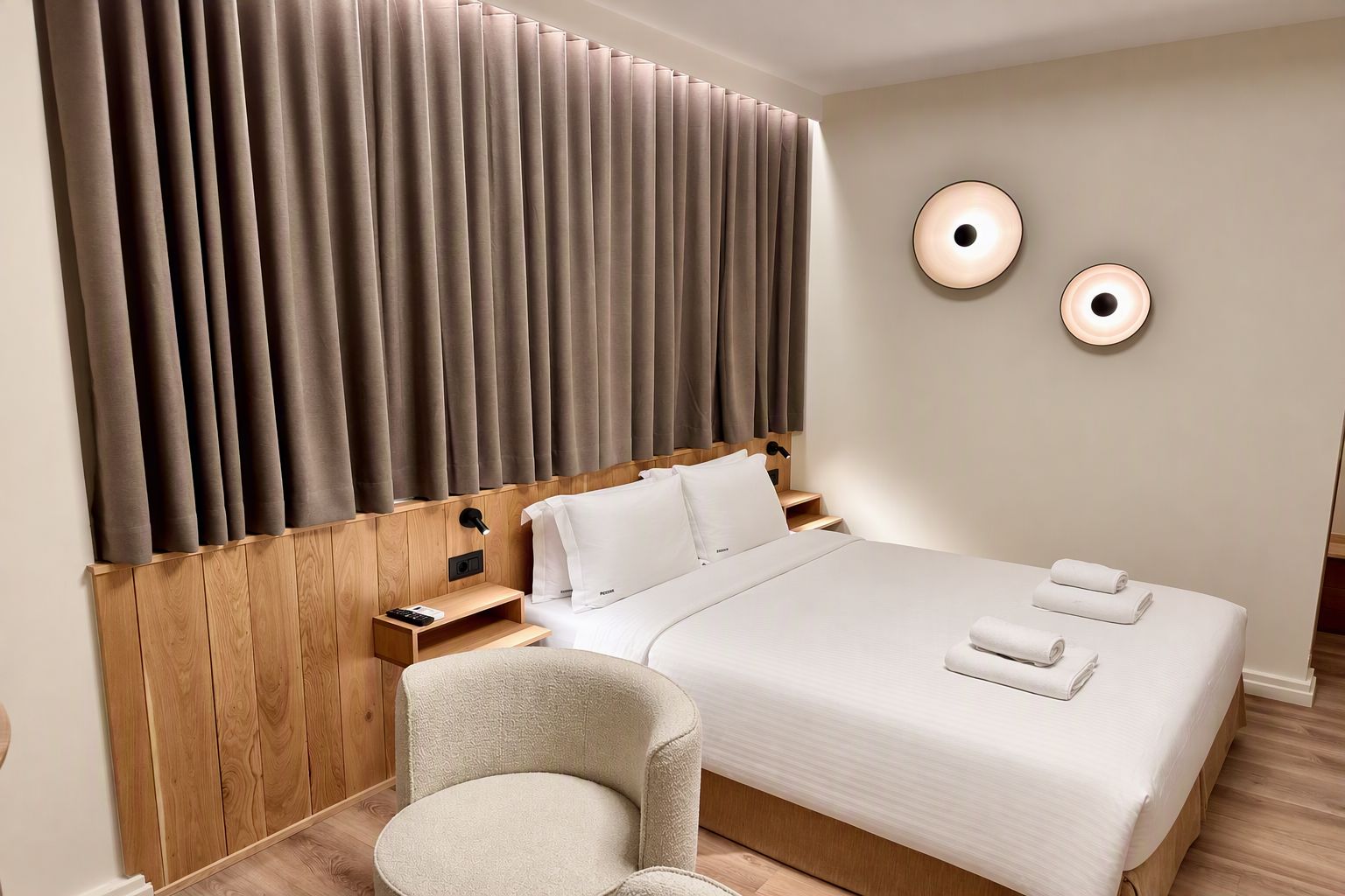 Une chambre d’hôtel avec un grand lit, une tête de lit en bois, une table de chevet, une chaise et deux lampes fixées au mur.