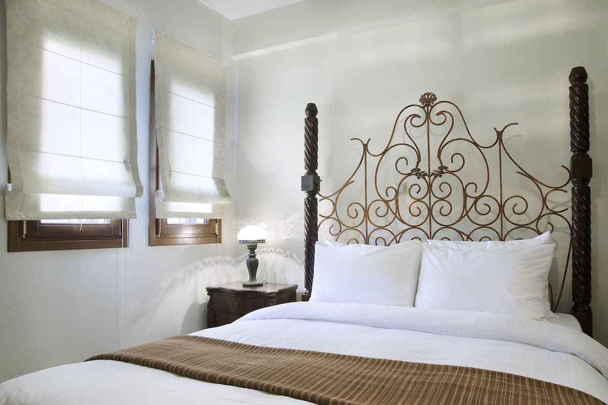 Un lit bien fait, recouvert d’une couverture marron à rayures, avec une tête de lit en métal ornée de motifs complexes, une table de chevet avec une lampe et une fenêtre équipée de stores.