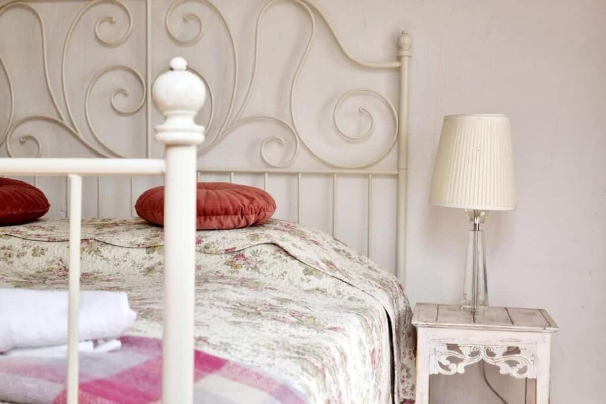 A bed with a floral bedspread, a white metal headboard, red pillows, and a white bedside table with a lamp.