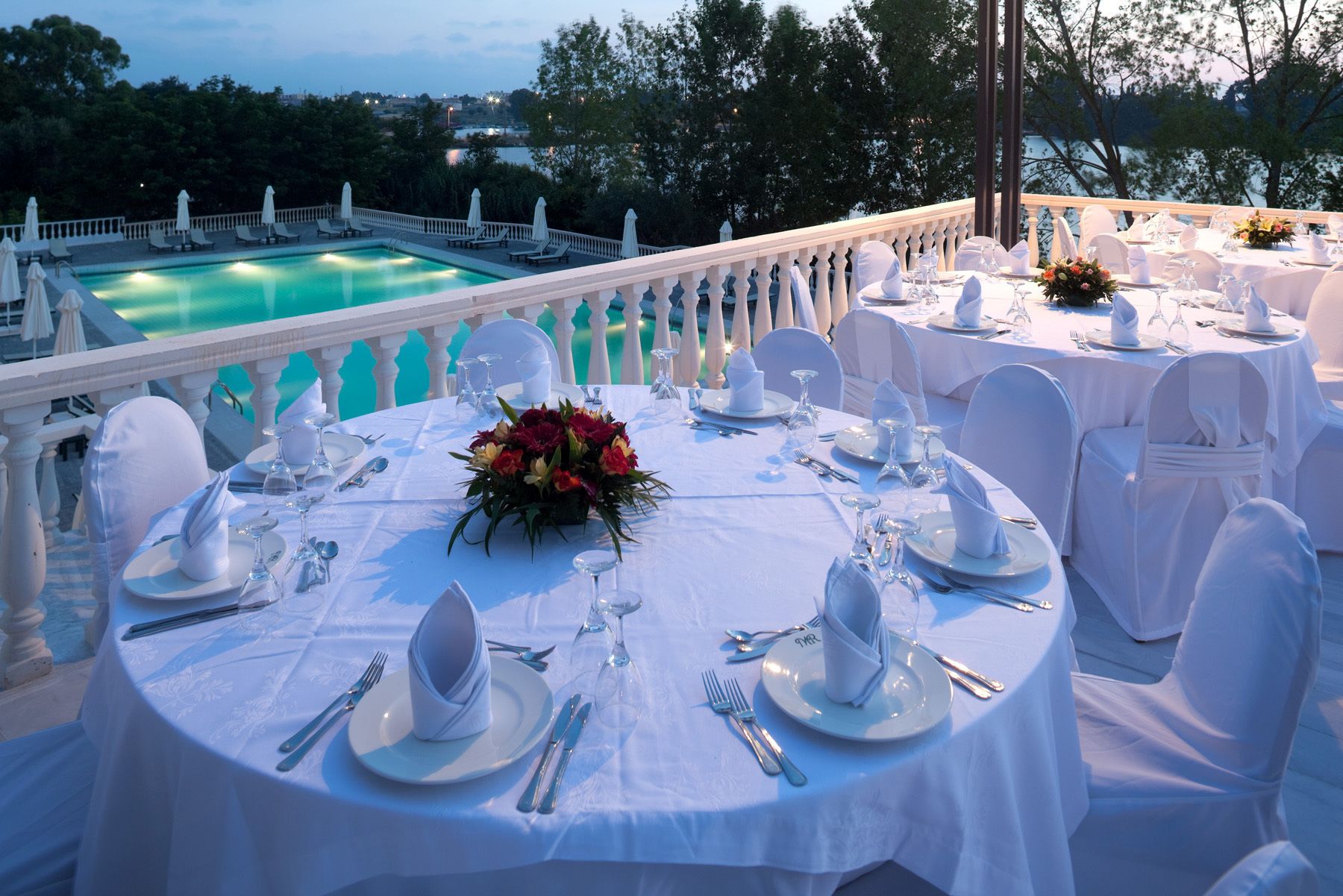 Un área de comedor exterior formal con mesas redondas dispuestas para una comida, manteles blancos y una piscina al fondo.