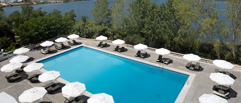 A rectangular swimming pool with a clear blue water, surrounded by lounge chairs and white umbrellas, overlooking a body of water and a distant town.