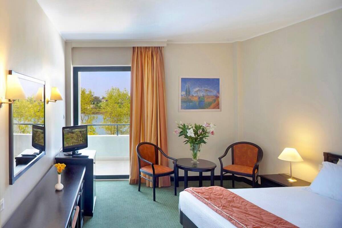 A hotel room with a bed, a desk with a computer, two chairs, a small table with flowers, a painting on the wall, and a view of trees and a bridge outside the window.